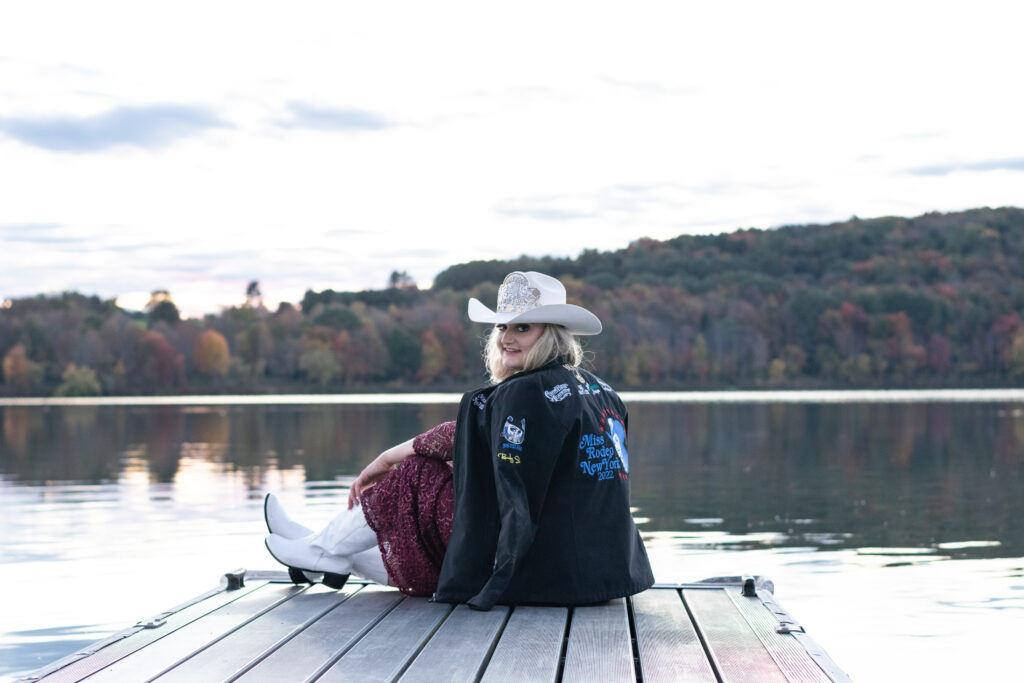 Miss Rodeo NY poses sitting at the end of a dock, showing off her sponsor jacket.